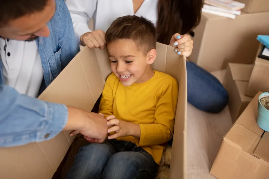 Young child smiling while sitting in a cardboard box as parents pack belongings nearby, suggesting a family move or transition.