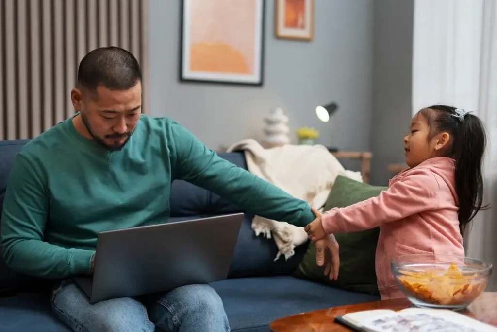 Parent working on a laptop at home while a young child reaches for attention, reflecting the balance between work responsibilities and parenting.