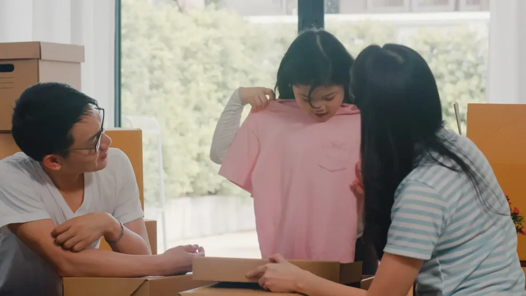 Parents and child unpacking boxes together in a new home, with the child holding up a shirt, reflecting family transition and shared responsibilities.