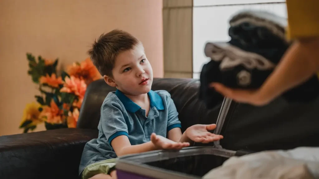 Young child sitting near an open suitcase while a parent packs clothes, suggesting family travel or transition between homes.