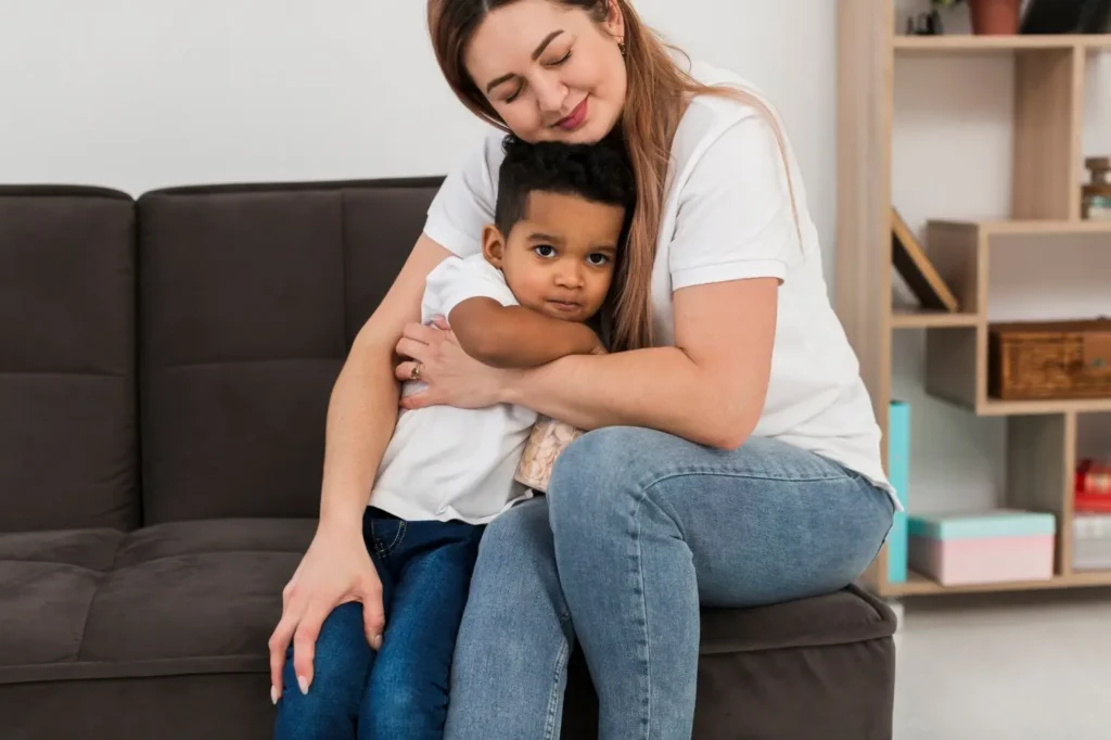 Parent sitting on a couch hugging a young child, expressing comfort, reassurance, and a strong emotional bond.
