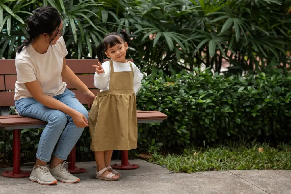 Parent sitting on a park bench beside a young child who gestures playfully, reflecting attentive supervision and a positive parent-child interaction outdoors.