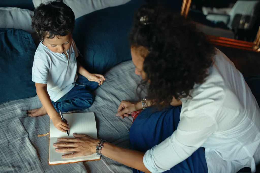 Mother helping young child practice writing in notebook on bed