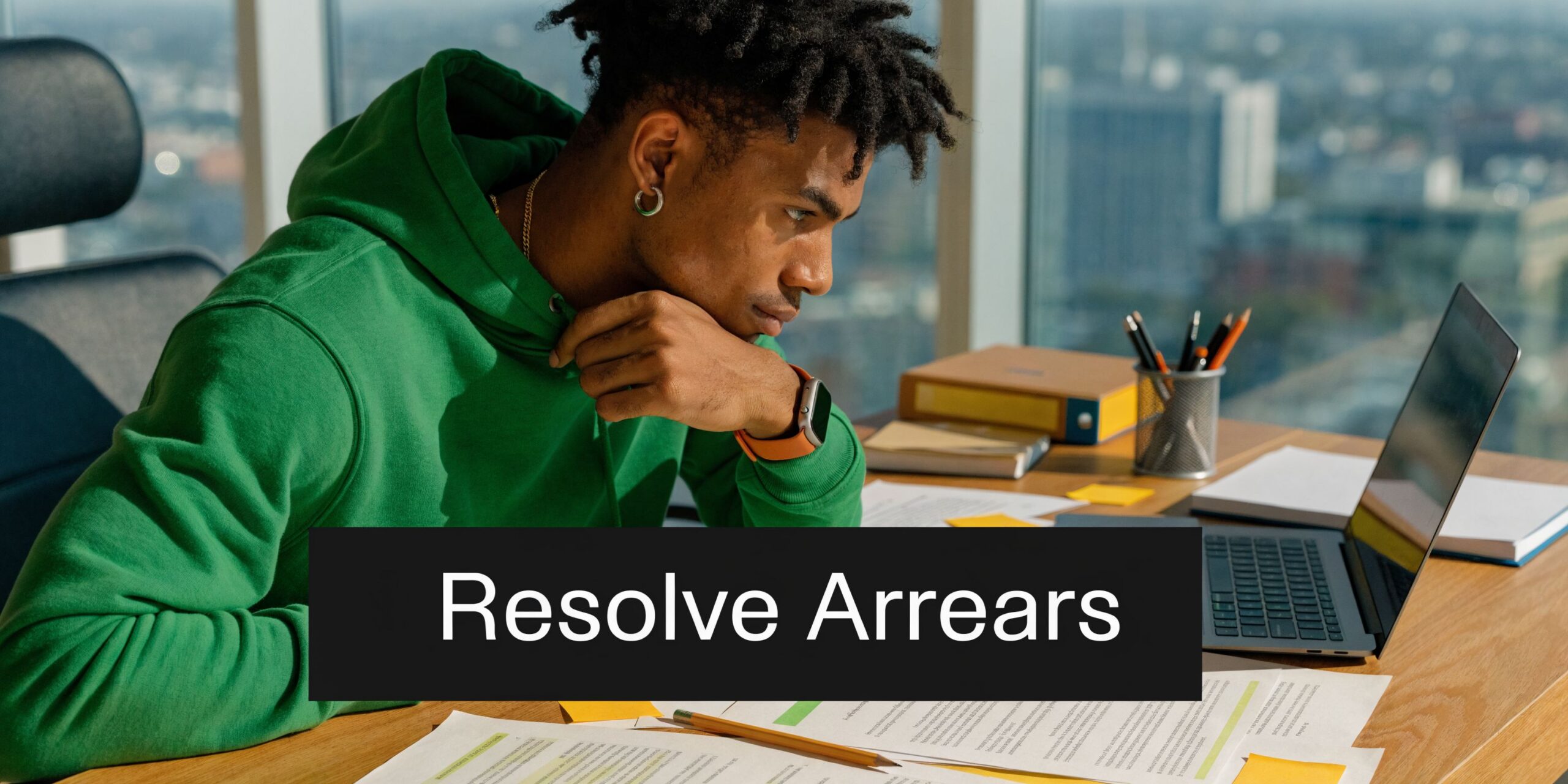 A young man in a green hoodie focused on his laptop while reviewing legal documents at a desk.