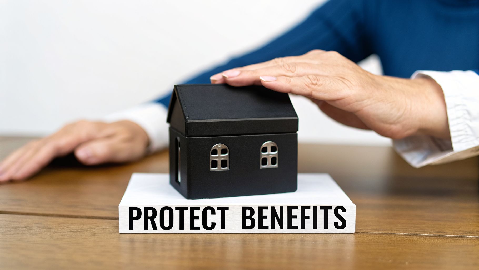 A person's hand gently covers a black miniature house on a block saying 'PROTECT BENEFITS'.